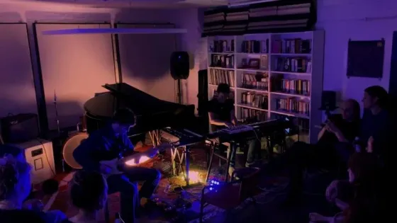 Fausto Cassarà and Tinkley playing live in the studio, in purple light, with an audience around them.